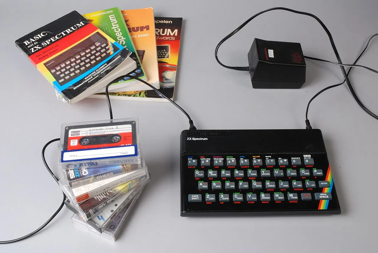 ZX Spectrum 48K із гумовою клавіатурою й блоком живлення; поряд — стос програмних і ігрових касет та кілька книжок про Spectrum.