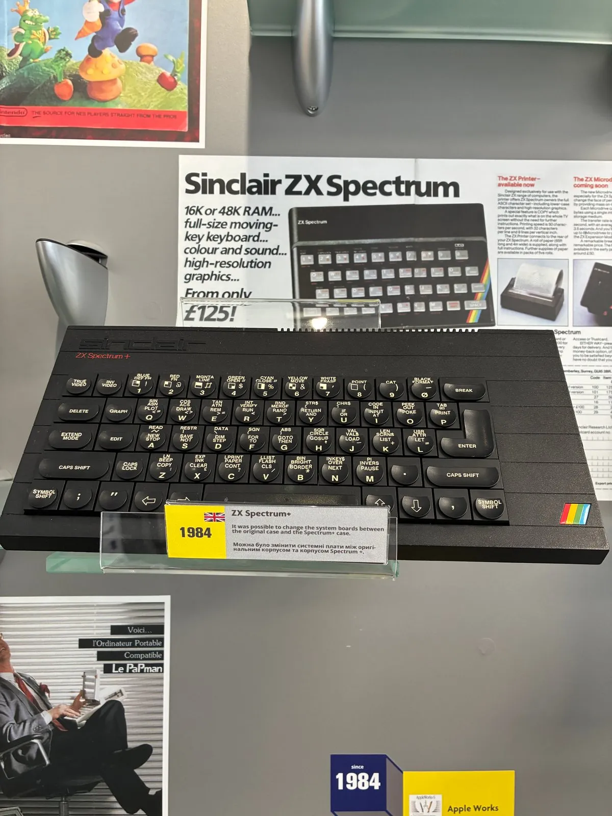 ZX Spectrum+ у вітрині музею: чорний корпус із повнорозмірною клавіатурою та райдужною смугою, поряд музейна табличка «1984» і рекламний плакат Sinclair